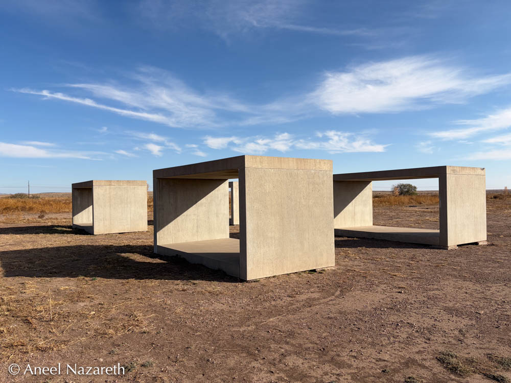 Four large concrete boxes grouped together, one of the 15 untitled works in concrete by Donald Judd. All of the boxes in the larger piece are 1 unit wide and tall and 2 units long. This set of four all have floor, celiling, and short walls in place, but the long walls are not present. They're grouped in a 2 x 2 rectangle, but the fourth box can barely be seen in this photo.